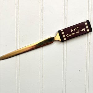 May include: A vintage letter opener with a gold-colored blade and a dark maroon handle. The handle is inscribed with "AHS Class of '49" in gold lettering. The letter opener is set against a white background.