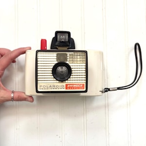 May include: Vintage Polaroid Swinger Model 20 camera. The camera is off-white with a black viewfinder, lens, and strap. The text "POLAROID LAND CAMERA SWINGER MODEL 20" is printed on the front in black and red. A red button is on top.