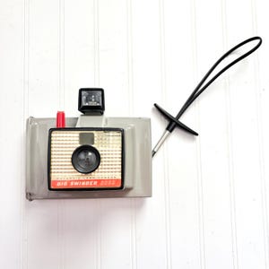 May include: A vintage Polaroid Big Swinger 3000 camera. The camera is gray with a black lens, flash, and strap. The front of the camera has a red and white label with the words "Big Swinger 3000".