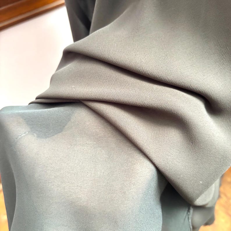 May include: Close-up of folded fabric in shades of gray and olive green. The fabric appears to be lightweight and slightly sheer, with visible folds and textures. The lighting highlights the different tones and the drape of the material.