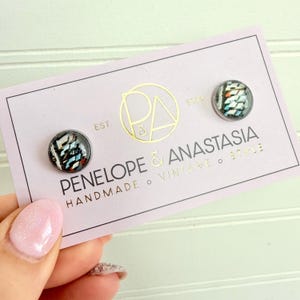 May include: A pair of round stud earrings with a colorful abstract design, displayed on a light pink card. The card features gold foil text that reads "PENELOPE & ANASTASIA HANDMADE VINTAGE STYLE". The earrings have a black background with blue, white, and red accents.
