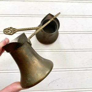 May include: Two antique brass Turkish coffee pots with a decorative handle and a matching spoon. The pots are made of a hammered metal and have a traditional design.