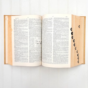 May include: An open, vintage dictionary with aged, yellowed pages. The book is bound in a light brown cover, with black tabs along the side for easy navigation. The pages are filled with small text and illustrations.