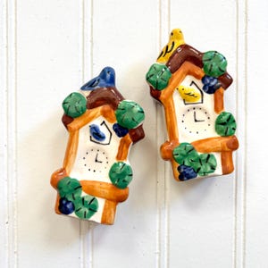 May include: Two ceramic cuckoo clock ornaments with a brown roof, white clock face, and yellow and blue birds. The ornaments are decorated with green leaves and brown branches.