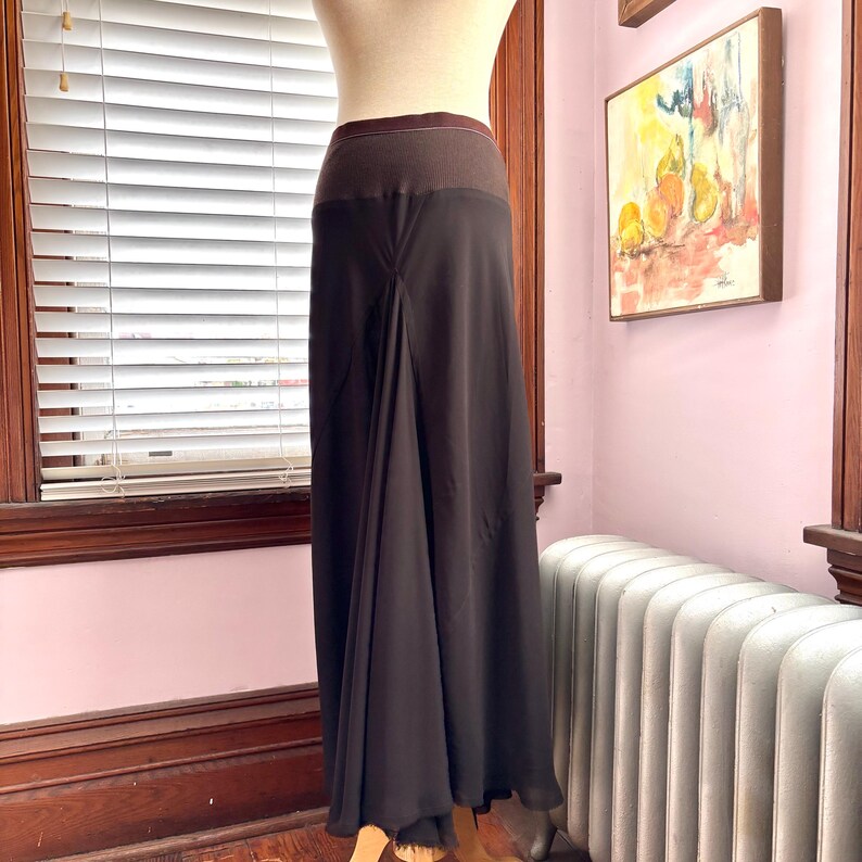 May include: A dark brown maxi skirt with a wide, dark brown waistband. The skirt has a flowing, A-line silhouette and is displayed on a mannequin. The skirt is made of a lightweight material.