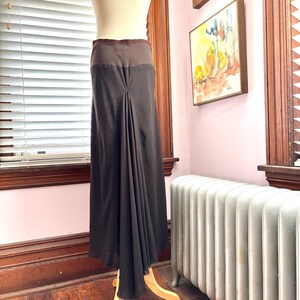 May include: A dark brown maxi skirt with a contrasting brown waistband is displayed on a mannequin. The skirt features a draped design that creates a flowing, elegant silhouette. The skirt is made of a lightweight fabric.