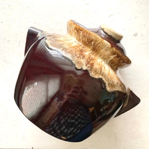 May include: A brown and cream glazed ceramic crock pot with a lid. The crock pot has a unique, irregular shape with a rounded bottom and a wide opening.