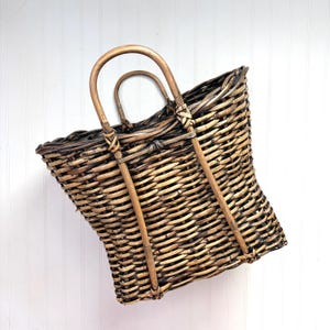 May include: A woven wicker basket with two arched handles. The basket is made of light brown and dark brown wicker, with a rectangular shape and a slightly curved design. The handles are made of a lighter-colored wood and are attached to the basket with woven details.