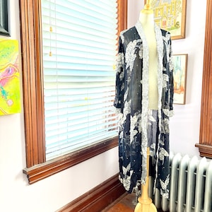 May include: A long, open-front duster cardigan in sheer black fabric featuring a white floral pattern and silver sequin embellishments. The garment has long sleeves and a wide, textured silver sequin trim along the front opening and cuffs.
