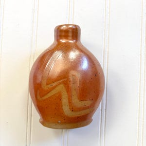 May include: A small, brown ceramic bottle with a rounded body and a narrow neck. The bottle features a light tan, zigzag design on the front. The surface has a speckled texture. The bottle is likely used for storing liquids.
