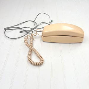 Peut inclure: Un téléphone beige vintage avec un cordon de combiné enroulé et un cordon d'alimentation droit. Le téléphone a une base rectangulaire avec un dessus incurvé. Les cordons sont gris et beiges, sur un fond blanc.