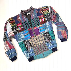May include: A long-sleeve zip-up jacket featuring a patchwork design composed of various woven fabrics in shades of blue, purple, red, green, and yellow. The garment includes a stand-up collar, ribbed cuffs, a ribbed hem, and two front pockets.