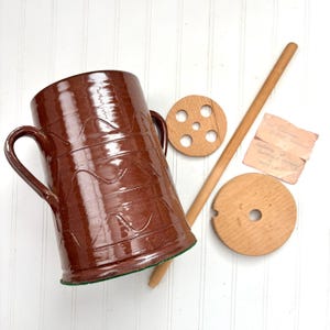 May include: A brown ceramic butter churn with two handles and a decorative wave pattern. Includes wooden components: a lid, a perforated disc, a long stirring stick, and a small handwritten note. The churn is approximately 10 inches tall.