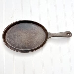 May include: Oval cast iron skillet with a single handle. The pan is dark gray with a well-seasoned surface. The handle has a hole for hanging. The skillet is designed for stovetop or oven use.