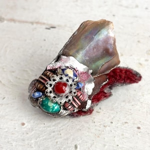 May include: A unique, handcrafted brooch featuring a central red gemstone surrounded by a silver filigree design. The brooch is adorned with various beads and set on a piece of iridescent shell and red fabric.