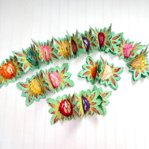 May include: A colorful paper garland featuring accordion-style flowers in shades of orange, pink, yellow, and purple. The flowers are attached to green leaf-shaped cutouts with red and yellow accents. The garland is displayed on a white surface.