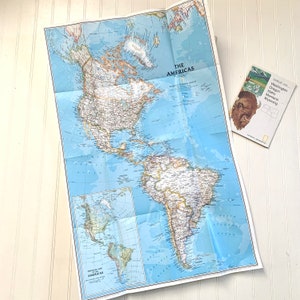 May include: A folded map of The Americas, showing North and South America, with a blue background and brown lines for borders and rivers. The map is titled "The Americas" and has a smaller inset map of Central America. The map is on a white surface.