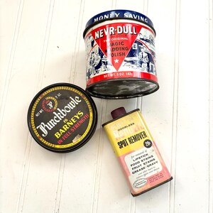 May include: Three vintage tins on a white surface. The tins include a black and gold Punchbowle tin, a red, white, and blue Nevr-Dull tin, and a yellow and pink Spot Remover tin. The Nevr-Dull tin reads "Money Saving".