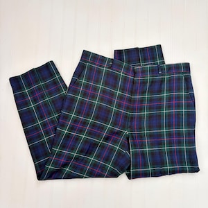 May include: A pair of plaid pants in a classic tartan pattern. The pants feature a navy blue, green, black, red, and white color scheme. The pants are folded and laid flat on a white surface.
