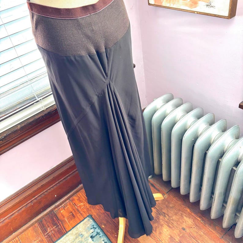 May include: A dark gray skirt with a brown ribbed waistband and a flowing, asymmetrical design. The skirt is displayed on a mannequin, showcasing its draping and texture. The skirt's fabric appears to be lightweight.