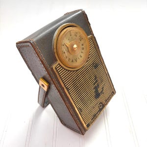 Puede incluir: Radio transistor RCA Victor Deluxe vintage en una funda de cuero gris. La radio tiene un dial y una rejilla de altavoz dorados. La funda tiene un pequeño soporte de cuero y herrajes dorados.