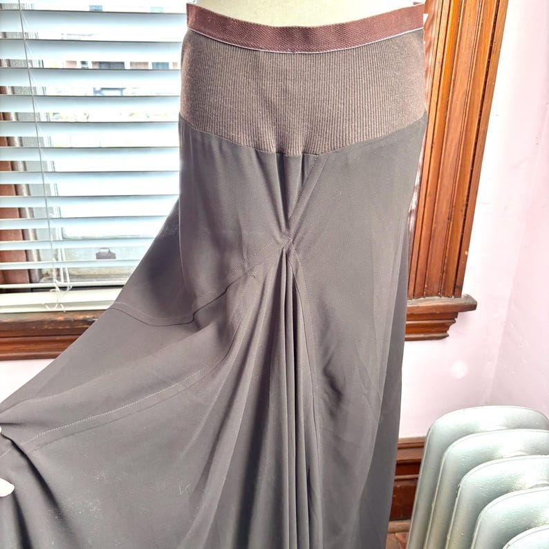 May include: A dark brown skirt with a ribbed knit waistband and a flowing, pleated design. The skirt has a subtle sheen and a flared silhouette, suitable for various occasions.