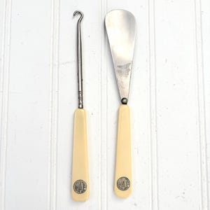 May include: Two vintage ivory-colored tools with metal components. One is a shoe horn, the other a hook-shaped tool. Both have ivory handles with a circular emblem. The tools are set against a white, vertical-slat background.