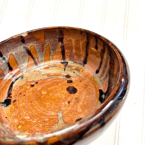 May include: A handmade ceramic bowl with a glossy finish. The bowl features a blend of warm colors, including shades of brown, orange, and tan, with dark brown and black accents. The interior has a textured, swirling pattern.