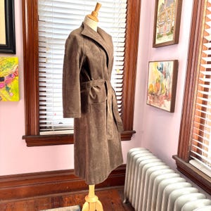 May include: A brown suede coat with a belted waist and two front pockets. The coat is displayed on a wooden mannequin in front of a window with blinds. The coat has a long length and a lapel collar.