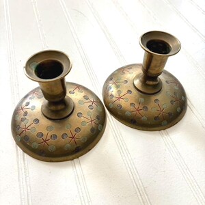 May include: Two vintage brass candle holders. Each holder has a round base with a decorative pattern of red and green starbursts and dots. The candle holders have a short, cylindrical stem and a circular cup to hold a candle.