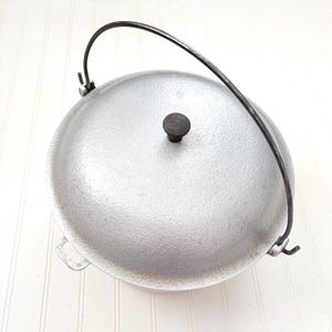 May include: A silver-colored, round cooking pot with a lid and a black metal handle. The pot has a textured surface and a small, dark knob on the lid. The handle arches over the top, attached to the sides.