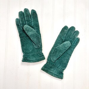 May include: A pair of dark forest green gloves featuring a soft, textured suede-like finish. Each glove has a gathered elastic wrist detail and five fingers, designed with a simple pull-on style that covers the hand and wrist area completely.