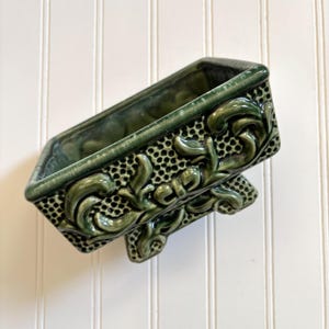 以下が含まれることがあります： 複雑な花柄と4本の脚が付いた緑色のセラミック製のプランター。プランターは正方形で、表面はテクスチャ加工されています。