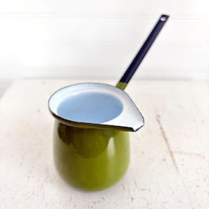 May include: An olive green enamel milk pan with a white interior and a long, dark blue handle. The pan features a wide, flat pouring spout on one side and a smooth, glossy finish on the exterior surface, designed for heating liquids on a stovetop.