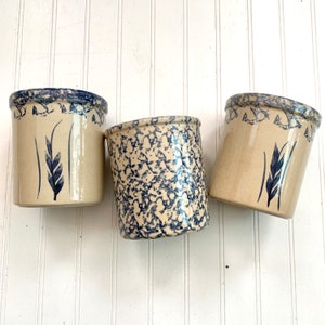 May include: Three ceramic crocks with blue and white speckled glaze. Each crock has a blue wheat design on the front.