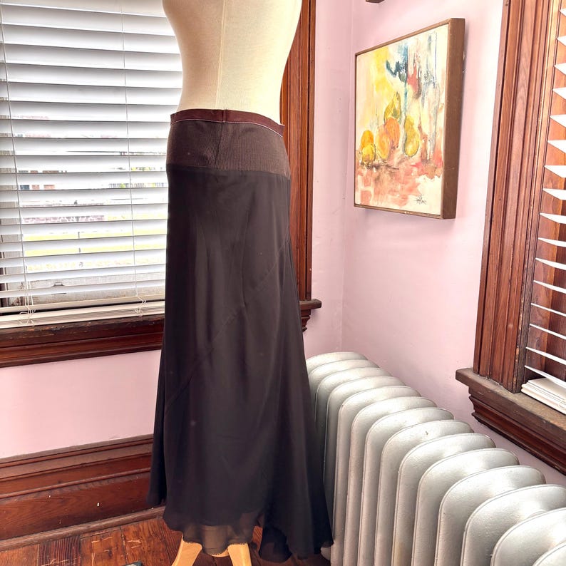 May include: A long, dark brown skirt displayed on a mannequin. The skirt has a wide, textured waistband and a flowing, sheer lower section. The skirt is floor-length and appears to be made of a lightweight material.