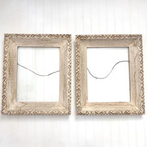 May include: Two rectangular picture frames with a light beige, weathered wood finish. Each frame features an ornate, carved border pattern along the outer edge and a thin, dark wire stretched horizontally across the center for hanging items.