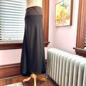 May include: A long, dark brown skirt on a mannequin. The skirt has a wide, textured brown waistband and a flowing, sheer lower section. The hemline features a black trim. The skirt is displayed on a wooden stand in a room with a window.