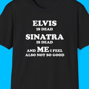 May include: Black t-shirt with white text that reads: "ELVIS IS DEAD, SINATRA IS DEAD, AND ME I FEEL ALSO NOT SO GOOD". The shirt has a crew neck and short sleeves. The background is a solid light blue.
