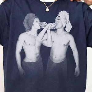 May include: Navy blue t-shirt with a black and white print of two people drinking from a can. The image shows the people wearing towels on their heads and around their waists. The t-shirt has a crew neck and short sleeves.