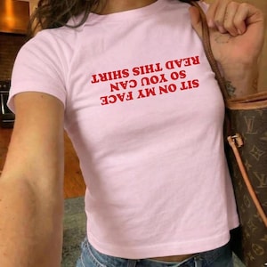 Peut inclure: T-shirt rose clair avec l'inscription rouge "SIT ON MY FACE SO YOU CAN READ THIS SHIRT". Le t-shirt est de style court et est porté avec un jean bleu.