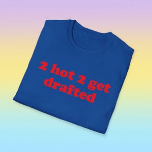 May include: A royal blue t-shirt with the phrase "2 hot 2 get drafted" in red, bold letters. The shirt is folded and presented on a gradient background of purple, yellow, and blue.