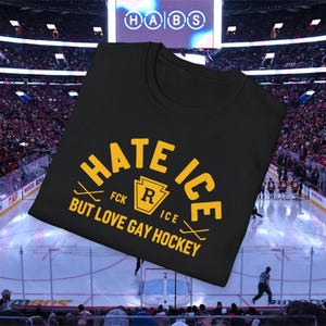 May include: Black t-shirt with the text "HATE ICE BUT LOVE GAY HOCKEY" in yellow. The design includes hockey sticks and a keystone shape with the letter "R" inside. The shirt is displayed in a hockey arena.