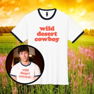 May include: White t-shirt with navy blue trim and the words "wild desert cowboy" in coral lettering. The image includes a circular inset of a person wearing the same shirt. The background features a field of pink flowers and a sunset.