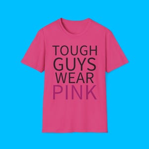Könnte beinhalten: Ein leuchtend pinkfarbenes T-Shirt mit dem Text "TOUGH GUYS WEAR PINK" in Schwarz und Lila. Das T-Shirt mit Rundhalsausschnitt wird vor einem blauen Hintergrund präsentiert. Der Text ist mittig auf der Vorderseite des Kleidungsstücks platziert.