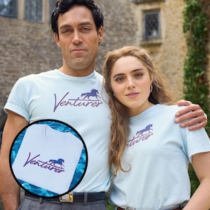 以下が含まれることがあります： 水色のTシャツを着た2人。各シャツには、紫色のスクリプトで「Venturer」という単語と、紫色の馬のグラフィックが描かれています。Tシャツは半袖です。背景には石造りの建物があります。