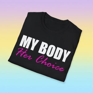 Op de afbeelding: Zwart T-shirt met de tekst "MY BODY" in het wit en "Her Choice" in het roze. Het shirt is opgevouwen en wordt weergegeven op een gradiënt achtergrond van geel, blauw en paars.