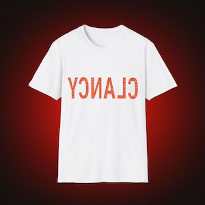 May include: A white short-sleeved t-shirt featuring the word CLANCY printed in large, bold, orange capital letters across the chest. The text is oriented upside down, creating a distinct visual contrast against the plain white fabric of the shirt.