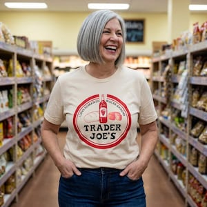 May include: A woman wearing a beige t-shirt with a red and white circular graphic. The graphic includes the text "Trader Joe's" and illustrations of a wine bottle, bread, and cheese. The woman is smiling in a grocery store.