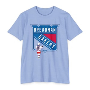 May include: Light blue t-shirt featuring a graphic of a hockey player and a logo that reads "Breadman Bakery".